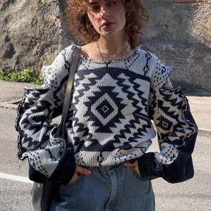 May include: A black and white patterned sweater with cut-out shoulders and long sleeves. The sweater features a geometric design and is paired with blue jeans. The sleeves have a dark denim fabric.
