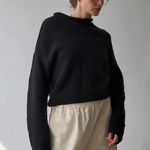 以下が含まれることがあります： 黒色のハイネック長袖セーターと、明るいベージュ色のパンツの組み合わせ。セーターはテクスチャード加工が施されています。モデルはゴールドフレームの眼鏡をかけ、髪を後ろにまとめています。