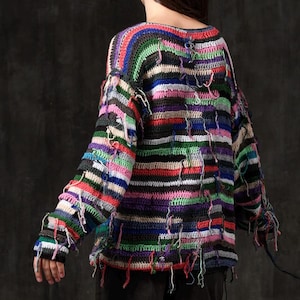 Könnte beinhalten: Ein langärmeliger Häkelpullover mit horizontalen Streifen in verschiedenen Farben wie Schwarz, Weiß, Rot, Blau, Grün und Pink. Das Kleidungsstück hat eine lockere, strukturierte Strickoptik mit zahlreichen losen Garnenden, die überall herabhängen.