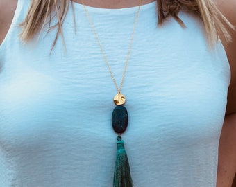 Puede incluir: Un collar de cadena dorada con un colgante ovalado verde oscuro y un disco dorado. El colgante tiene un borla de hilo verde oscuro.