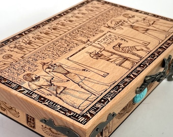 May include: A wooden box with an intricate design of Egyptian hieroglyphs and figures. The box has metal hinges and feet, and a turquoise stone accent.