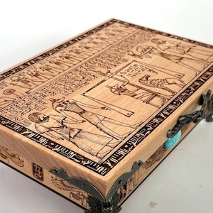 Puede incluir: Una caja de madera con un diseño intrincado de jeroglíficos y figuras egipcios. La caja tiene bisagras y patas de metal, y un acento de piedra turquesa.