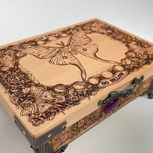 May include: A wooden box with a hinged lid and ornate metal accents. The lid features a woodburned design of a luna moth with a moon phase pattern and the text "AS ABOVE SO BELOW".
