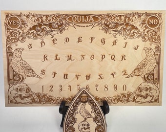 Può includere: Un tabellone Ouija in legno con una finitura colorata marrone. Il tabellone presenta una disposizione tradizionale con lettere, numeri e le parole "YES" e "NO". Il tabellone è decorato con un design in stile gotico con corvi e teschi.