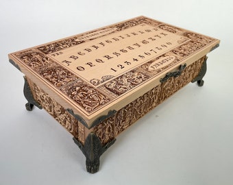 May include: A wooden box with a hinged lid and ornate carvings. The lid features a wooden planchette with an alphabet, numbers, and the words "YES" and "NO". The box has four decorative metal feet.
