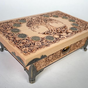 May include: A wooden box with a Celtic tree of life design and a metal latch. The box has a light wood finish and is decorated with intricate carvings. The text "As Above So Below" is engraved on the top of the box.
