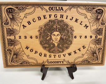 May include: A wooden Ouija board with engraved letters, numbers, and a central illustration of a woman with snakes for hair. The board includes the words "YES", "NO", "OUIJA", and "GOODBYE". The board is displayed on a dark brown stand.
