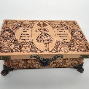 Puede incluir: Una caja de madera con tapa con bisagras y detalles metálicos. La tapa está decorada con un diseño intrincado que presenta a Alicia de Alicia en el País de las Maravillas, relojes y la cita "No sirve de nada volver al ayer, porque yo era una persona diferente entonces."