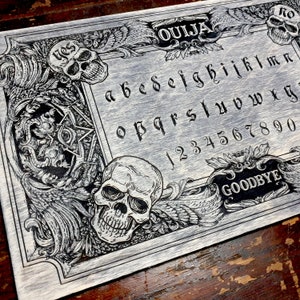May include: A black and white wooden Ouija board with a skull design. The board features the letters of the alphabet, numbers 1-9 and 0, and the words "Ouija", "Goodbye", and "Yes".