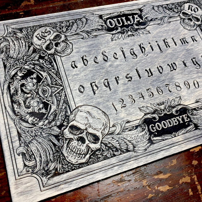 May include: A black and white wooden Ouija board with a skull design. The board features the letters of the alphabet, numbers 1-9 and 0, and the words "Ouija", "Goodbye", and "Yes".