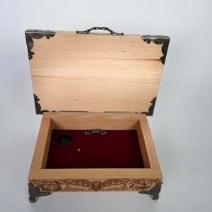 May include: A wooden music box with a red velvet interior and intricate carvings on the exterior. The box has a hinged lid with a metal handle and decorative metal corners.