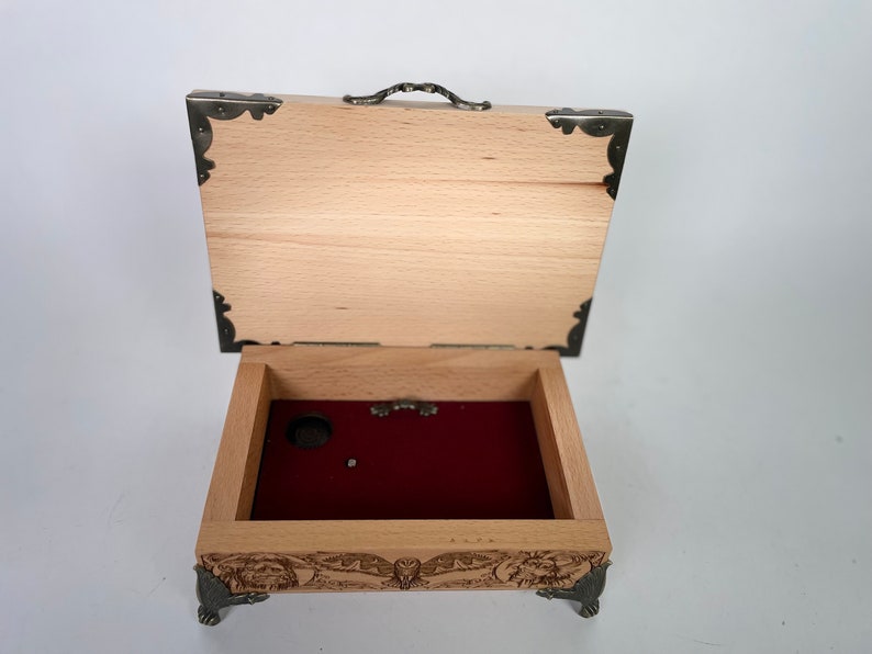 May include: A wooden music box with a red velvet interior and intricate carvings on the exterior. The box has a hinged lid with a metal handle and decorative metal corners.