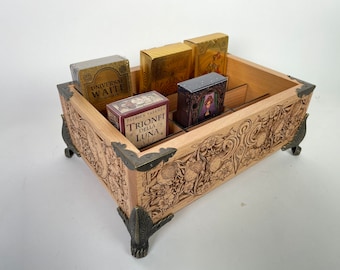 May include: A wooden box with intricate carvings and brass accents, designed to hold and display multiple decks of tarot cards. The box has four compartments and is filled with various tarot decks, including "Universal Waite" and "Trionfi della Luna".