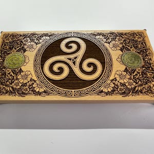Op de afbeelding: Een rechthoekige houten kist met een Keltisch ontwerp. De kist heeft een centrale donkerbruine cirkel met een drievoudige spiraalknoop, omgeven door een lichtere houten rand met bloemen- en knoopwerk-snijwerk. Twee cirkelvormige messingkleurige accenten bevinden zich aan de zijkanten.