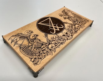 May include: A rectangular wooden box with a detailed laser-etched design featuring skeletons, skulls, and a sigil. The box has decorative metal feet. The artwork is in shades of brown and black.