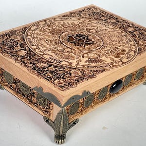 Op de afbeelding: Een houten doos met ingewikkelde houtsnijwerk van een cirkel met een pentagram in het midden. De doos heeft sierlijke metalen poten en een zwarte steen aan de zijkant.