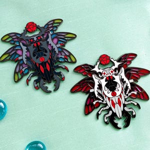 May include: Two insect-themed enamel pins with intricate wing designs, one black and the other white with red accents, rest on a light green fabric stretched within a wooden embroidery hoop. Blue glass beads and floral accents complete the composition.