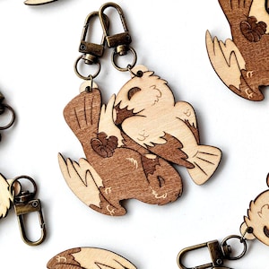 Könnte beinhalten: Zwei hölzerne Eulenschlüsselanhänger, in Braun und Beige bemalt, miteinander verschlungen. Die Schlüsselanhänger haben einen goldenen Metallverschluss.