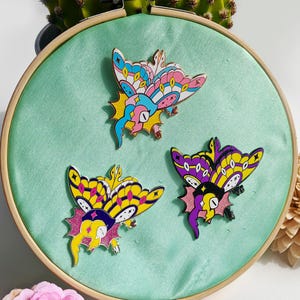 Op de afbeelding: Drie emaillepins met kleurrijke vlinderontwerpen. De vlinders zijn in verschillende tinten roze, geel, paars en blauw. De pins worden getoond op een groene stoffen achtergrond.