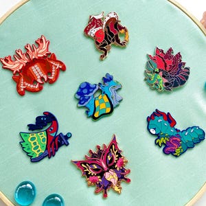 May include: A collection of colorful enamel pins with various designs, including a red octopus, a red and black creature, a blue and yellow mushroom, and a floral mask. The pins are arranged on a light green fabric stretched within a wooden embroidery hoop.
