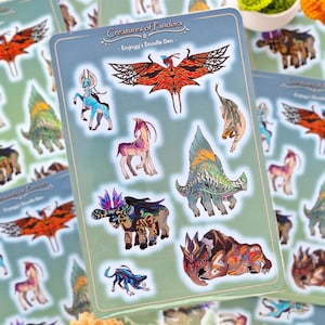 Puede incluir: Hojas de pegatinas con criaturas fantásticas de colores. Las pegatinas están sobre un fondo azul claro con el texto "Creatures of Pandora - Erujoyy's Doodle Den -" en la parte superior. Las pegatinas representan varias criaturas fantásticas.