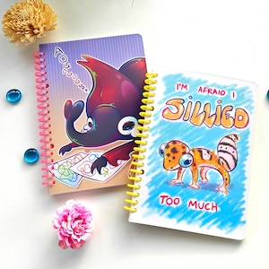 May include: Two spiral bound notebooks with colorful covers. The first notebook has a purple cover with a cartoon illustration of a red and black bug with the text "POYOJOJOJO" on the cover. The second notebook has a blue cover with a cartoon illustration of a yellow and white lizard with the text "I'M AFRAID I SILLIED TOO MUCH" on the cover.