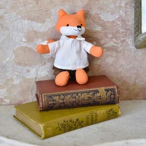 May include: A plush fox toy with orange fur, white face and chest, and brown shorts. The fox is wearing a white collared shirt. It is standing on top of two antique books with gold lettering.