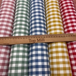 Pode incluir: Vários rolos de tecido em várias cores, incluindo vermelho, verde, azul e amarelo, cada um com um padrão xadrez clássico. Uma régua de madeira com o texto "Top Mill" é colocada sobre os rolos de tecido, com medidas em cm e mm.