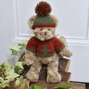 May include: A plush teddy bear wearing a knitted red sweater with a green Christmas tree design and a matching green and red hat with a pom-pom. The bear is light brown and sitting on a wooden surface.