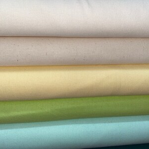 May include: Five rolls of fabric in different colors. The top roll is a light beige, the second roll is a slightly darker beige, the third roll is a pale yellow, the fourth roll is a light green, and the bottom roll is a light blue.