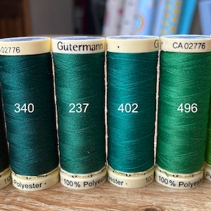 Puede incluir: Seis carretes de hilo verde y verde lima, cada uno con una etiqueta blanca. Las etiquetas tienen el texto "Gütermann" y un número en cada carrete. Los números son 472, 340, 237, 402, 496 y 336.