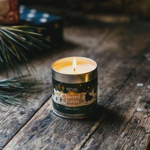 May include: A lit candle in a silver tin with the words "Sacred Forest" printed on the label. The candle sits on a rustic wooden surface. The label features a forest scene with a dark green and gold color scheme.