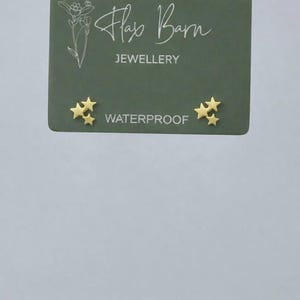 May include: Gold-colored star-shaped stud earrings. The earrings are displayed on a green card with the text "Flax Barn Jewellery" and "Waterproof". The earrings are small and delicate.