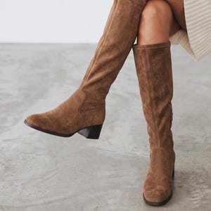 May include: A pair of knee-high brown suede boots featuring a rounded toe, a low block heel, and a vertical seam running down the center of the shaft. The boots have a smooth, matte texture and a simple, clean design that reaches just below the knee.