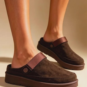 Può includere: Un paio di zoccoli in camoscio marrone scuro con una spessa suola platform in gomma in tinta e un ampio cinturino in tessuto regolabile sulla parte superiore, fissato con un bottone circolare sul lato di ogni scarpa per una calzata sicura.