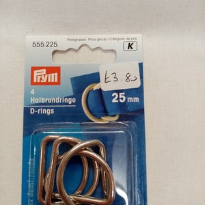 Könnte beinhalten: Packung mit vier silbernen D-Ringen, 25 mm groß, für Bastelprojekte. Die Verpackung ist blau und weiß mit dem Prym-Markenlogo und dem Text "Halbrundringe D-rings".