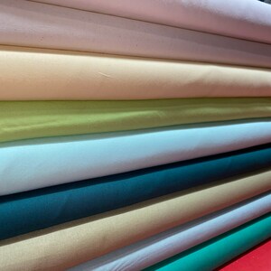 May include: A stack of fabric rolls in various colors, including white, cream, light green, light blue, teal, beige, and light blue. The fabric rolls are neatly stacked on top of each other.