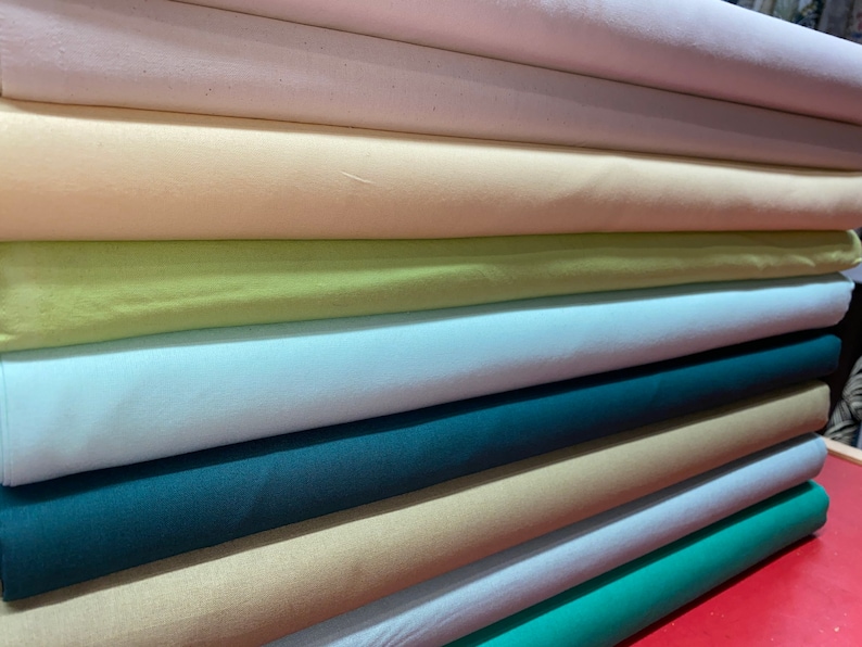 May include: A stack of fabric rolls in various colors, including white, cream, light green, light blue, teal, beige, and light blue. The fabric rolls are neatly stacked on top of each other.