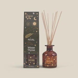 May include: A reed diffuser set featuring a dark green rectangular box with gold celestial illustrations, the text St. Eval and Winter Thyme, and a matching amber glass bottle with wooden reeds, also decorated with gold star patterns and the St. Eval logo.