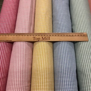 Puede incluir: Rollos de tela a cuadros en varios colores, incluyendo rojo, rosa, amarillo, azul y verde, con un patrón de cuadros clásico. Una regla de madera con el texto "Top Mill" se coloca sobre los rollos de tela. La regla mide hasta 30 centímetros.