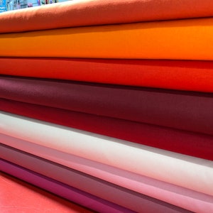 May include: A stack of eight fabric rolls in various shades of orange, red, and pink. The fabric is smooth and appears to be cotton.