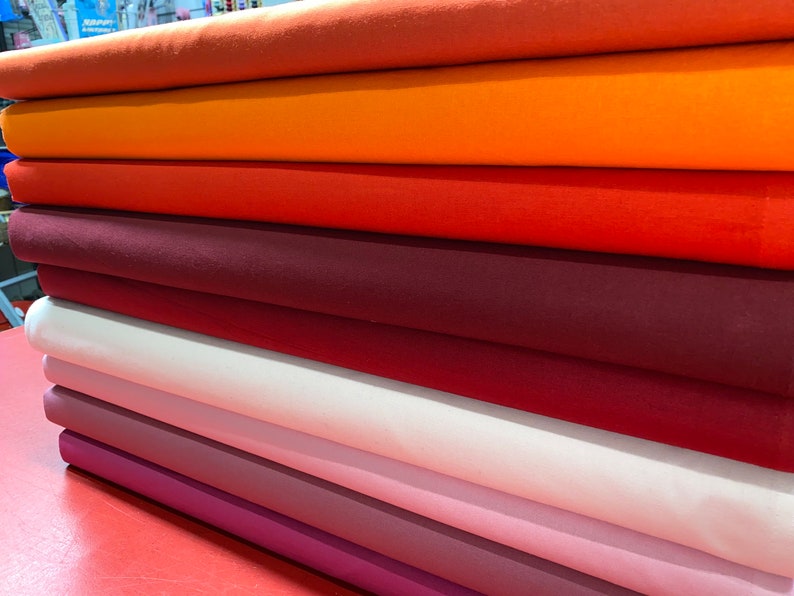 May include: A stack of eight fabric rolls in various shades of orange, red, and pink. The fabric is smooth and appears to be cotton.