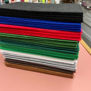 Op de afbeelding: Een stapel vellen vilt in verschillende kleuren, waaronder zwart, blauw, rood, groen, wit, grijs en bruin. De vellen vilt zijn netjes gerangschikt en tonen een reeks tinten die geschikt zijn voor knutsel- en doe-het-zelfprojecten.