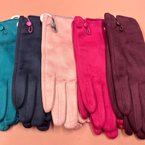 Könnte beinhalten: Ein Set aus fünf Paar wildlederartigen Handschuhen in den Farben Petrol, Marineblau, Hellrosa, Pink und Dunkelburgunder. Jedes Paar verfügt über einen kleinen Knopfverschluss am Handgelenk mit passender Schlaufe und ist nebeneinander angeordnet.