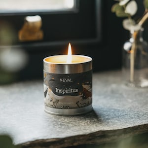 May include: A lit candle in a silver metal tin with a dark label featuring the text St. Eval and Inspiritus. The label displays a whimsical illustration of figures in a snowy landscape with stars. The candle flame glows brightly from the centre.