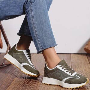Può includere: Sneakers verde oliva e bianche con un design strutturato. Le sneakers hanno suole e lacci bianchi e sono indossate con jeans blu. Le scarpe sono su un pavimento di legno.