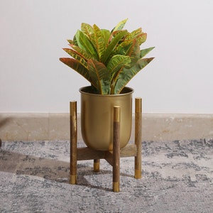 Op de afbeelding: Een gouden metalen plantenstandaard met een ronde bovenkant en drie poten. Een plant in een pot met groene en gele bladeren staat op de standaard.