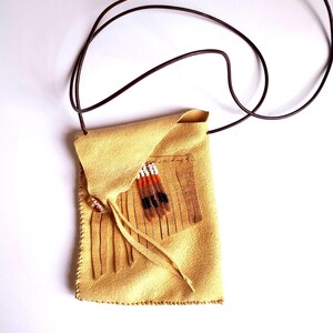 May include: A tan suede pouch with a brown leather cord. The pouch features a fringed pocket with white, orange, and black beaded accents. A button closure and fringe detail the front.