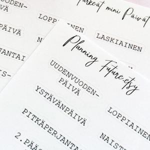 Op de afbeelding: Close-up van verschillende witte stickerbladen met Finse tekst, waaronder "Planning Future.etsy" in cursief. De stickers bevatten Finse feestdagen en data. Een decoratieve sneeuwvlok is zichtbaar op de achtergrond.