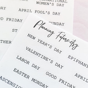 Op de afbeelding: Twee witte sticker vellen met zwarte tekst, met feestdagen zoals "Nieuwjaarsdag" en "Valentijnsdag". De vellen zijn gelabeld met "Planning Future.etsy" en "International Holidays". Decoratieve gouden sneeuwvlokken zijn op de achtergrond.